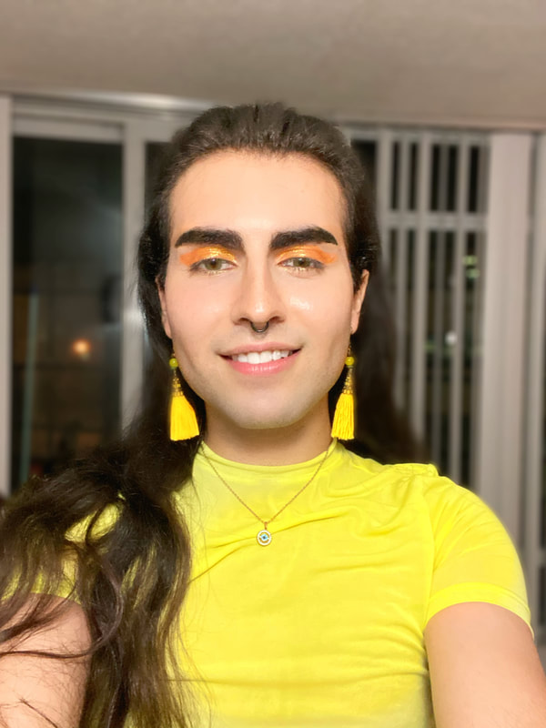 A photo of Arya (he/she/they) in their home. They are a Persian non-binary person with long dark brown hair. Arya is wearing a bright yellow shirt, matching yellow dangling earrings, a gold evil eye necklace...and a big smile!
