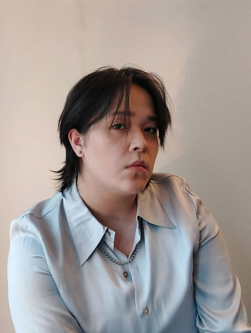 A photo of Sai Lian (he/him) against a tan background. He is a pale mixed asian man with his black hair in a mullet. Sai Lian is wearing a baby blue silk button-up shirt and silver jewelry.