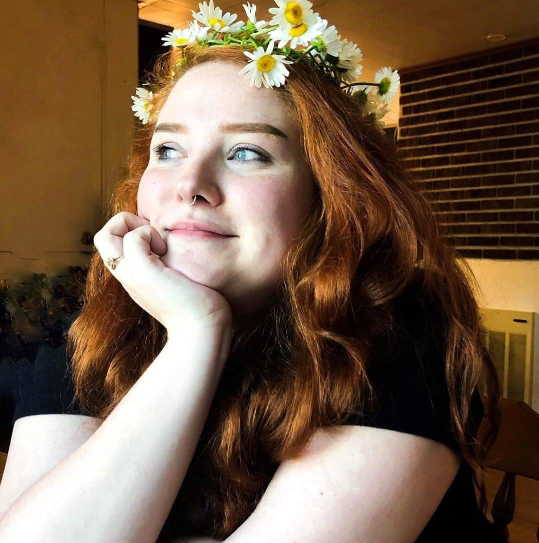  joey o’dael is a trans nonbinary person with long wavy red hair, straight thick brows, and blue eyes. They are resting their chin on their hand, as they smile slightly and look off-camera. They are wearing a braided daisy crown, small gold septum ring, and a black t-shirt.