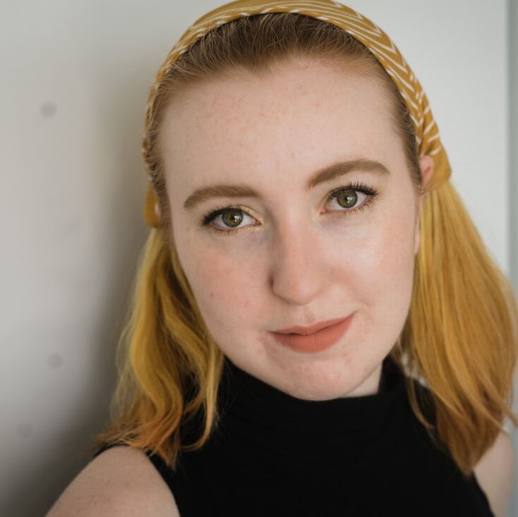 A picture of Katherine Teed-Arthur. She is a white woman in her mid-20s with red hair dyed yellow, wearing a black tank top and a yellow kerchief in her hair. She is smiling at the camera.